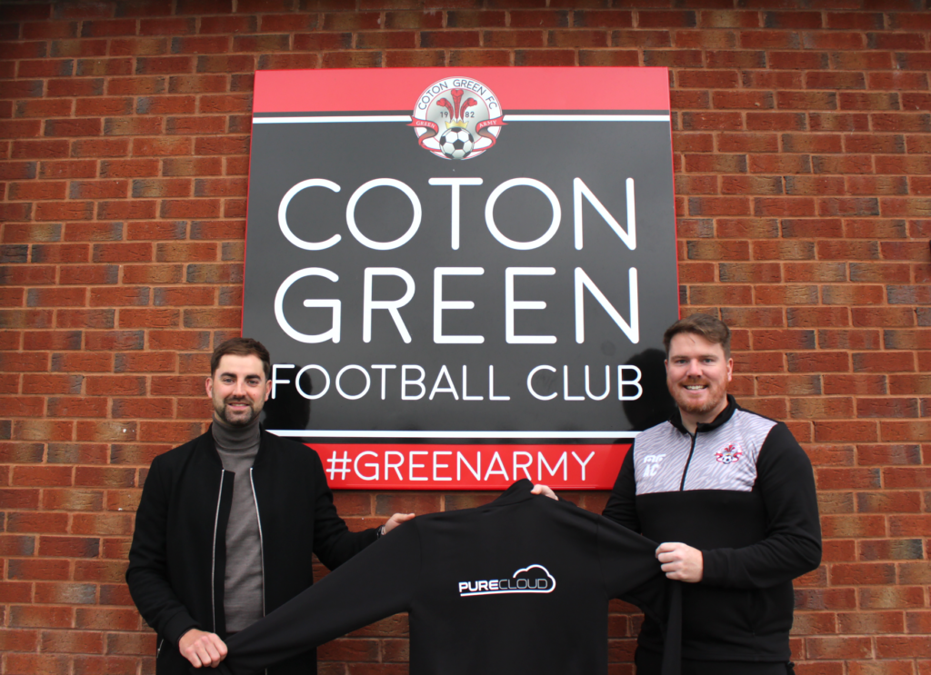 Two men holding a black sweatshirt with the PureCloud logo in front of the Coton Green Football Club sign.