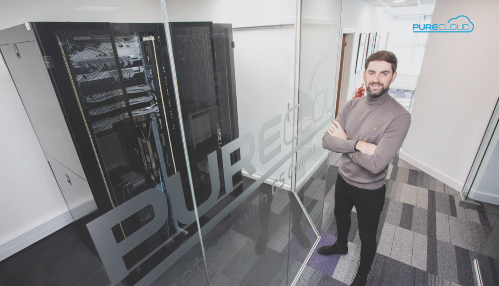 Jamie, Group CEO of PureCloud, standing confidently with arms crossed in front of a server rack in a modern office setting.