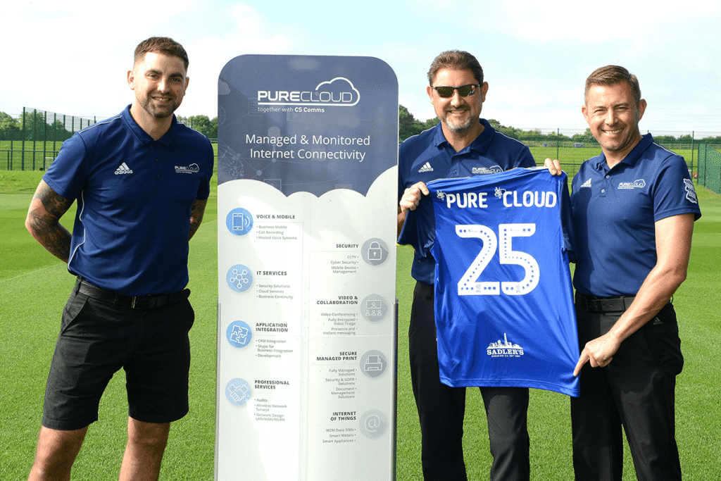 three men on a football field holding a jersey beside a pop-up banner